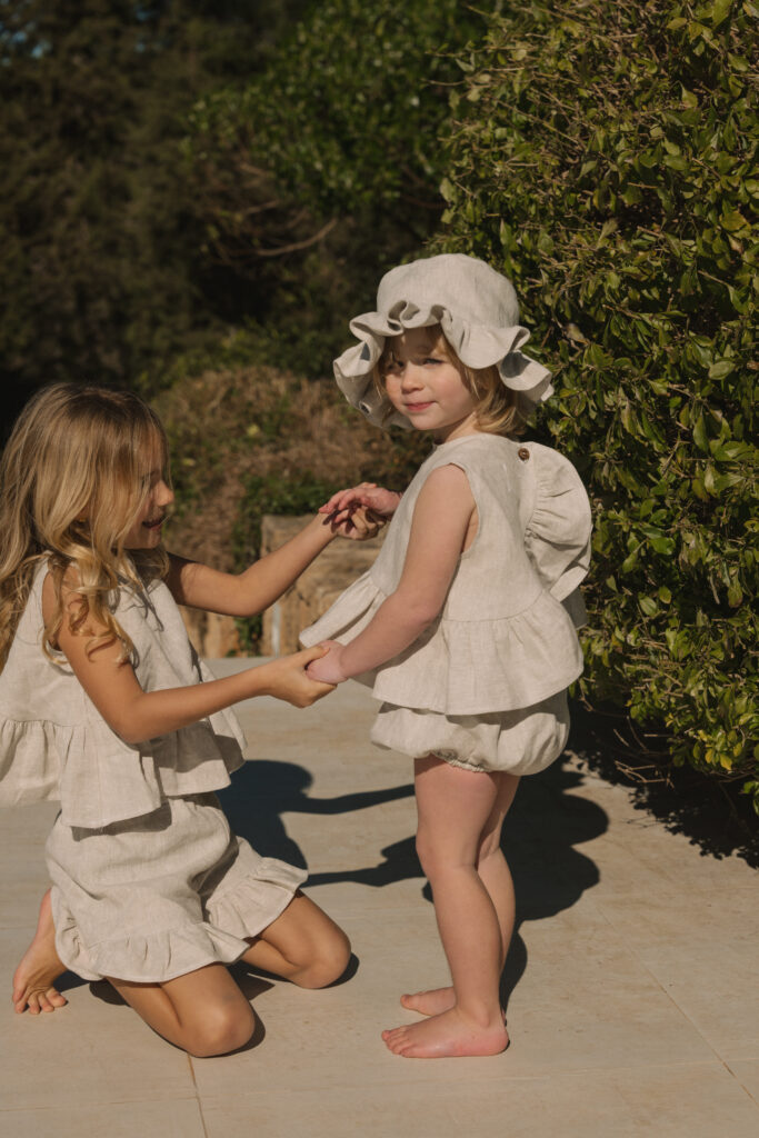 A baby girl in the sunshine at Summer wearing a linen frilled hat and matching frilled top and bloomers.