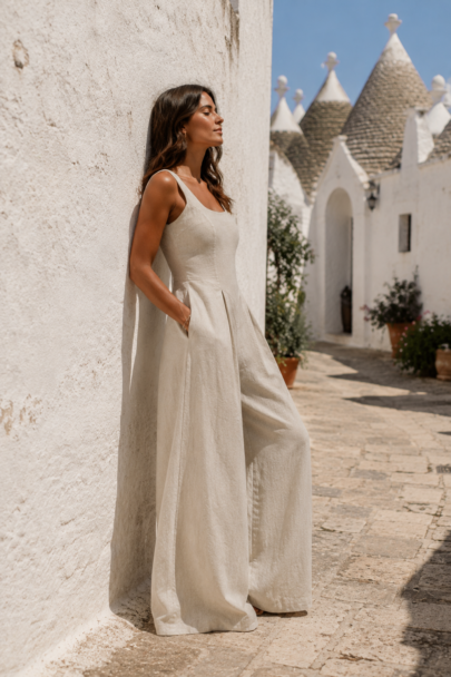 Glamourous tanned woman with long dark hair leaning against a white washed wall in Italy wearing a natural linen wide leg Freya Lillie jumpsuit.