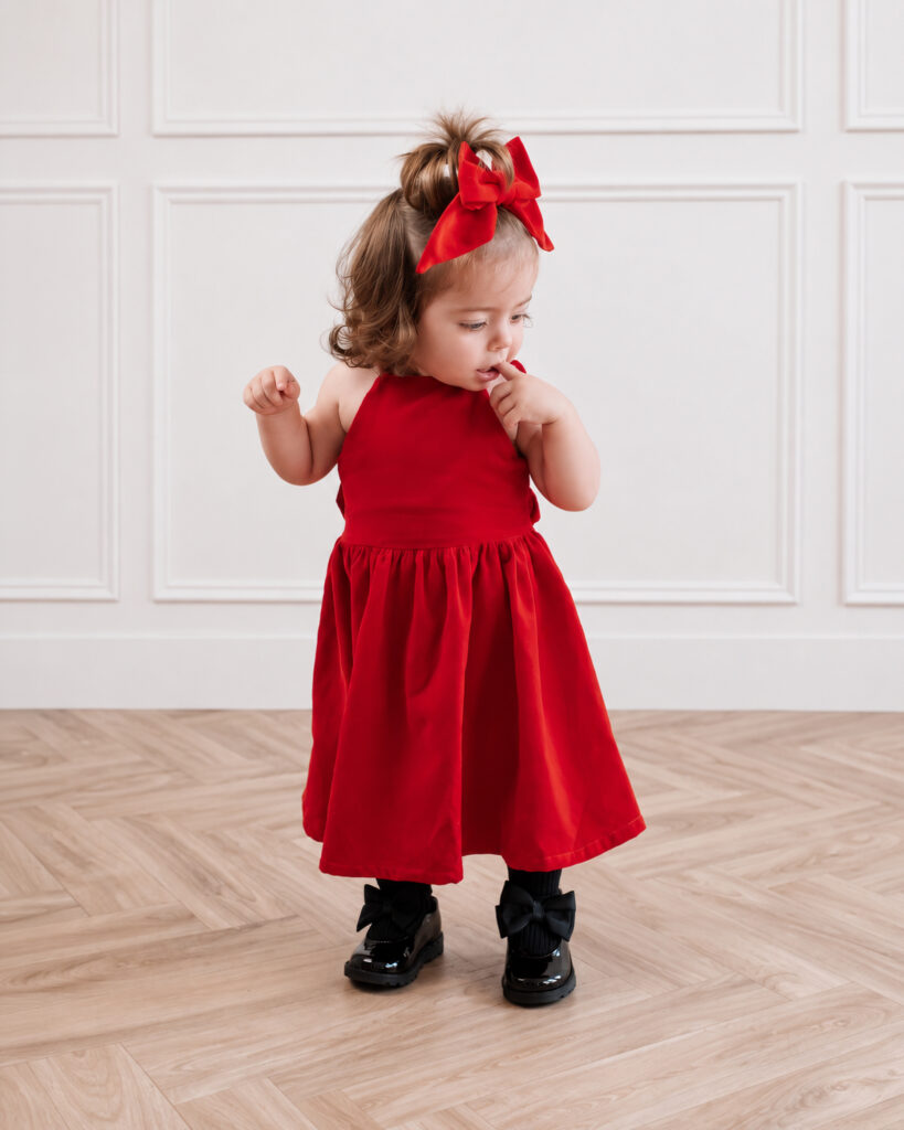 A toddler dark haired girl in a photo studio wearing a red velvet Freya Lillie Sorrento dress